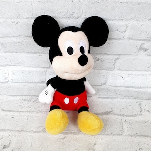 May include: A plush Mickey Mouse toy with black ears, a black nose, and white eyes. The toy is wearing red shorts with white polka dots and yellow shoes.