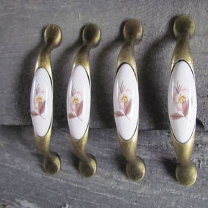 May include: Four antique brass cabinet knobs with white porcelain inserts featuring a floral design. The knobs have a curved shape and are ideal for adding a vintage touch to furniture.