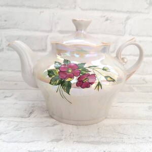 Pode incluir: Uma chaleira de porcelana branca com um design floral. A chaleira tem um acabamento perolado e apresenta flores rosa e verdes.