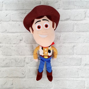 May include: A plush toy of Woody from Toy Story. He is wearing his signature brown hat, red bandana, yellow and brown plaid shirt, and blue jeans. He has a friendly smile and brown boots.