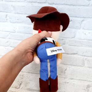 May include: A plush toy of Woody from Toy Story. He is wearing his signature brown hat, blue jeans, and a yellow and red plaid shirt. The toy has a brown belt and brown boots. The toy has a Disney Pixar tag on the back.