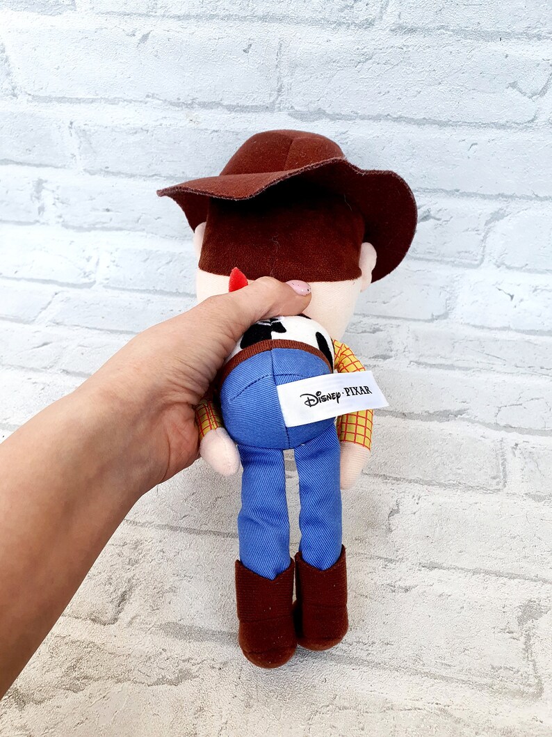 May include: A plush toy of Woody from Toy Story. He is wearing his signature brown hat, blue jeans, and a yellow and red plaid shirt. The toy has a brown belt and brown boots. The toy has a Disney Pixar tag on the back.
