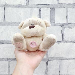 Könnte beinhalten: Ein beiger Teddybär mit brauner Nase und gestickten Details. Der Bär hat einen rosa Knopf am Bauch und einen kleinen Aufnäher am Fuß. Der Bär wird in einer Hand gehalten.
