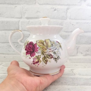Pode incluir: Bule de porcelana branca com um design floral com rosas vermelhas, folhas verdes e flores brancas. A chaleira tem uma borda dourada ao redor da borda e uma alça dourada.