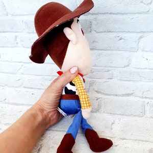 May include: A plush toy of Woody from the Toy Story franchise. Woody is wearing his signature brown cowboy hat, red bandana, yellow and blue plaid shirt, blue jeans, and brown boots. He is standing with his legs spread apart and his arms at his sides.