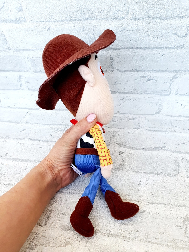 May include: A plush toy of Woody from the Toy Story franchise. Woody is wearing his signature brown cowboy hat, red bandana, yellow and blue plaid shirt, blue jeans, and brown boots. He is standing with his legs spread apart and his arms at his sides.