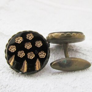 May include: A pair of cufflinks with a black background and gold floral pattern. The cufflinks are made of metal and have a rounded shape.
