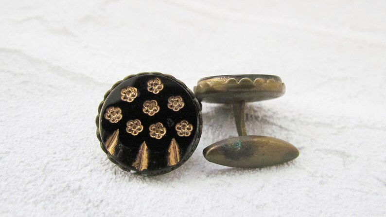 May include: A pair of cufflinks with a black background and gold floral pattern. The cufflinks are made of metal and have a rounded shape.