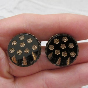 May include: A pair of round buttons in black and gold with a floral design. The buttons have a black background with gold flowers and a gold rim.