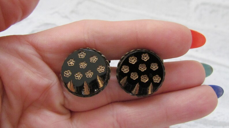 May include: A pair of round buttons in black and gold with a floral design. The buttons have a black background with gold flowers and a gold rim.