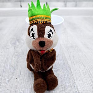 May include: A plush toy of a cartoon chipmunk wearing a green crown with gold trim. The chipmunk has brown fur, a white face, and a red tongue. The toy is standing on a light gray wooden surface.