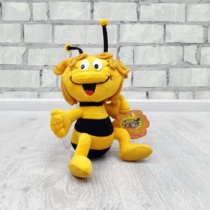 Könnte beinhalten: Ein Plüschtier einer Comic-Biene mit gelben und schwarzen Streifen. Die Biene hat ein lächelndes Gesicht, Fühler und ein Etikett mit der Aufschrift "Maya the Bee Friends". Das Spielzeug liegt auf einer hellen Holzoberfläche.