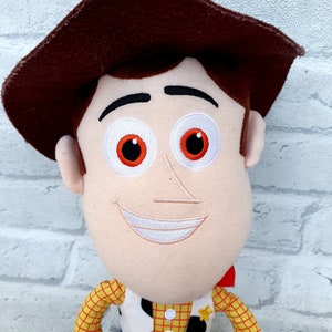 May include: A plush toy of Woody from the Toy Story franchise. Woody is wearing his signature brown hat, red and yellow plaid shirt, and blue jeans. He has a friendly smile and white eyes with orange pupils.