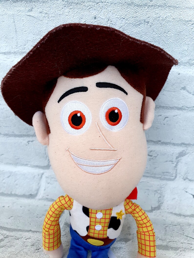 May include: A plush toy of Woody from the Toy Story franchise. Woody is wearing his signature brown hat, red and yellow plaid shirt, and blue jeans. He has a friendly smile and white eyes with orange pupils.