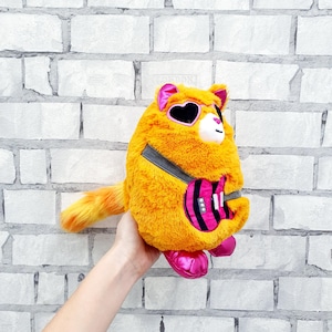 Pode incluir: Um brinquedo de pelúcia laranja em forma de gato com detalhes em rosa, usando óculos de sol em forma de coração e uma guitarra. O brinquedo tem uma alça cinza e pés rosa. A cauda do gato é laranja e amarela.