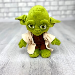 May include: Green plush Yoda toy wearing a brown robe and with a serious expression. The toy is standing on a light wood surface.