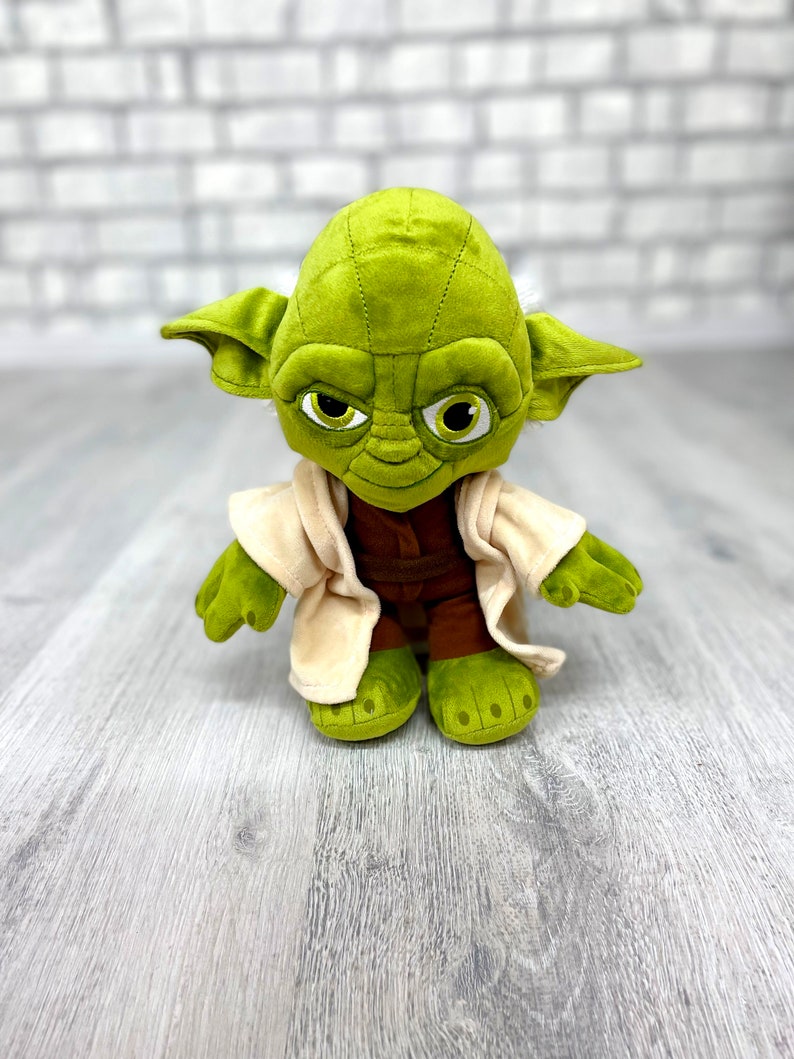 May include: Green plush Yoda toy wearing a brown robe and with a serious expression. The toy is standing on a light wood surface.
