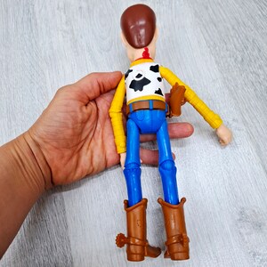 Puede incluir: Una figura de juguete de pl&aacute;stico de Woody de la franquicia Toy Story. Woody lleva su caracter&iacute;stico sombrero de vaquero marr&oacute;n, chaleco amarillo con bandana roja, pantalones vaqueros azules y botas de vaquero marrones. Est&aacute; extendiendo el brazo derecho, como si estuviera buscando algo.
