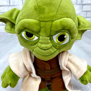 May include: Green plush Yoda toy wearing a tan robe. The toy has a brown belt and large, expressive eyes.
