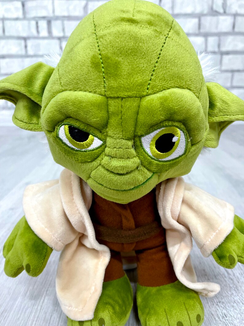 May include: Green plush Yoda toy wearing a tan robe. The toy has a brown belt and large, expressive eyes.
