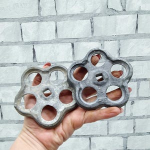 Op de afbeelding: Twee vintage, bloemvormige metalen kraanhendels in grijstinten. Elke hendel heeft een centraal vierkant gat en vijf cirkelvormige openingen. De hendels worden in een hand gehouden tegen een witte bakstenen muur achtergrond.