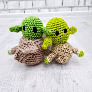May include: Two crocheted green Yoda characters with brown bodies. The Yoda on the left has a brown crocheted robe. The Yoda on the right has a brown crocheted robe with white accents.