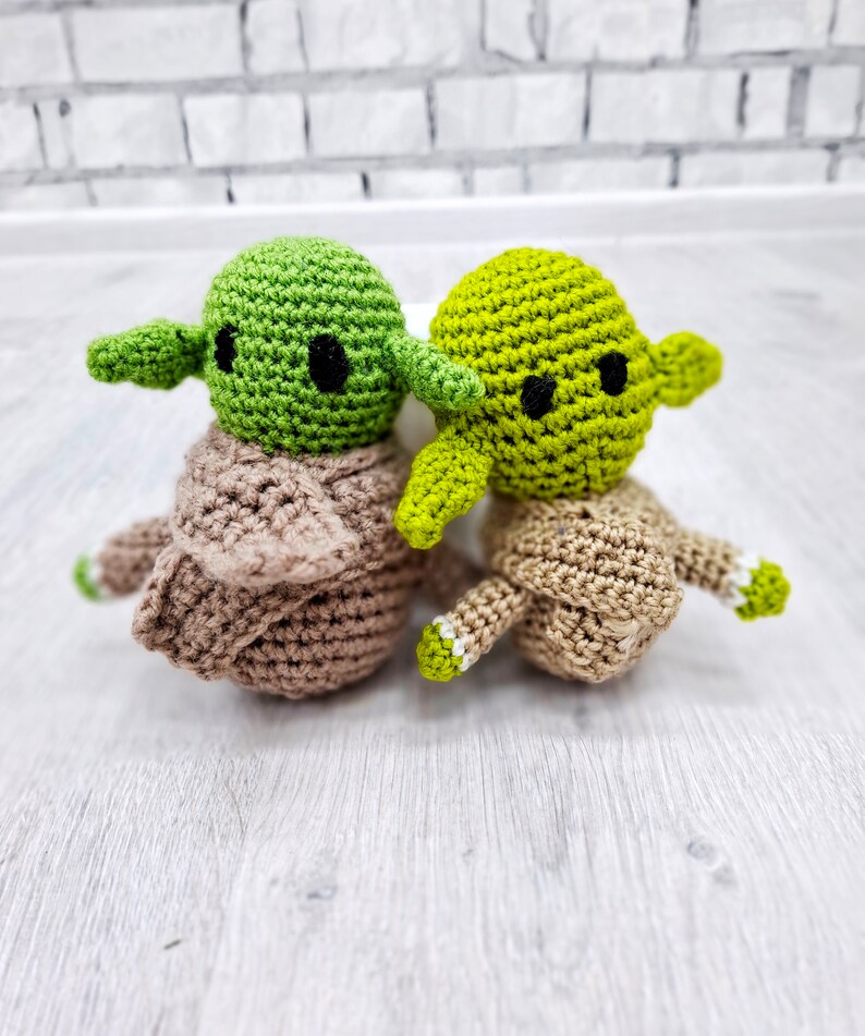 May include: Two crocheted green Yoda characters with brown bodies. The Yoda on the left has a brown crocheted robe. The Yoda on the right has a brown crocheted robe with white accents.
