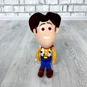 May include: A plush toy of Woody from Toy Story. He is wearing his signature brown cowboy hat, red bandana, and yellow vest with a sheriff's star. He has a friendly smile and brown eyes.