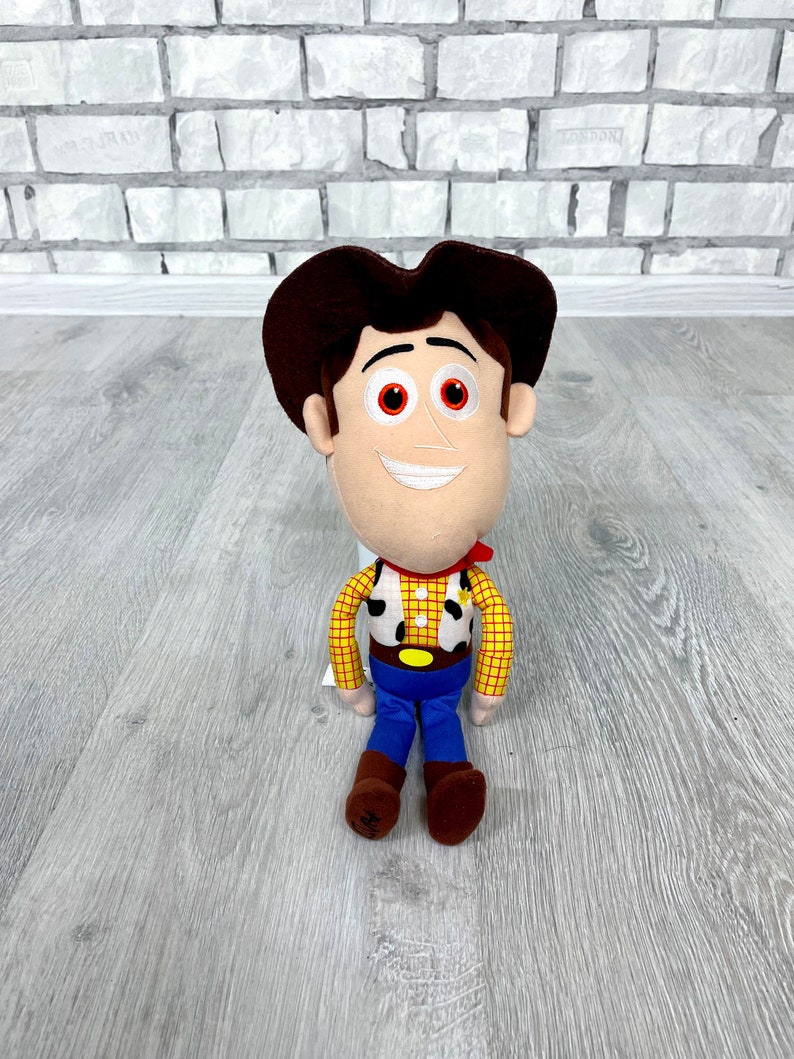May include: A plush toy of Woody from Toy Story. He is wearing his signature brown cowboy hat, red bandana, and yellow vest with a sheriff's star. He has a friendly smile and brown eyes.
