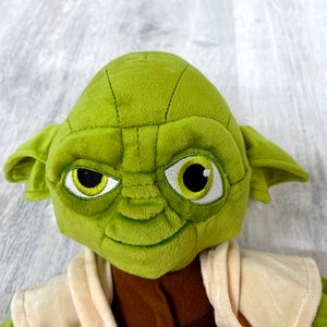 May include: Green plush Yoda toy with large ears, brown eyes, and a white robe.