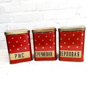 May include: Three red and gold metal canisters with white polka dots. The canisters are labeled "Rice", "Buckwheat", and "Pearl Barley" in Russian.