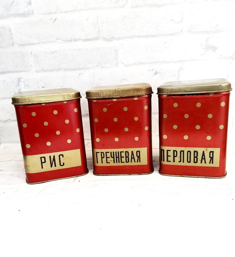 May include: Three red and gold metal canisters with white polka dots. The canisters are labeled "Rice", "Buckwheat", and "Pearl Barley" in Russian.