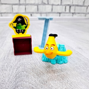 May include: A yellow Angry Birds character with a blue base and a blue and white stand. The character is wearing a pirate hat and holding a sword. The character is standing on a red box.
