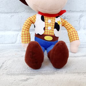 May include: A plush toy of Woody from Toy Story. He is wearing his signature cowboy hat, vest, and red bandana. He has a yellow star on his vest and brown boots.