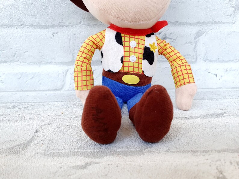May include: A plush toy of Woody from Toy Story. He is wearing his signature cowboy hat, vest, and red bandana. He has a yellow star on his vest and brown boots.
