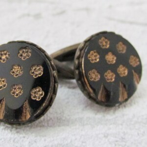May include: A pair of cufflinks with round black and gold buttons. The buttons feature a floral design.