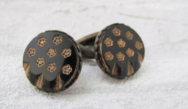 May include: A pair of cufflinks with round black and gold buttons. The buttons feature a floral design.
