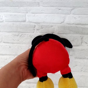 May include: A red and black plush toy shaped like Mickey Mouse. The toy is being held by a hand, and the back of the toy is facing the camera.