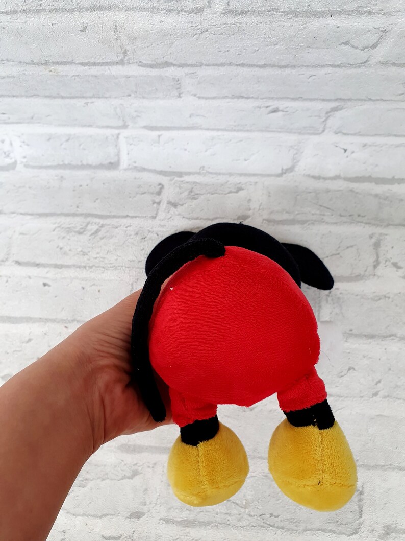 May include: A red and black plush toy shaped like Mickey Mouse. The toy is being held by a hand, and the back of the toy is facing the camera.