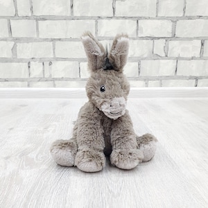 Peut inclure: Une peluche d'âne grise avec de grandes oreilles tombantes et un visage souriant. Le jouet en peluche est assis sur une surface en bois clair, avec un mur de briques blanches en arrière-plan. L'âne a une crinière et une queue touffues.