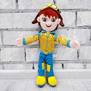 May include: A plush doll of a cartoon character wearing a yellow jacket with blue trim, blue pants with yellow patches, and yellow shoes. The doll has a brown aviator hat with goggles and red hair. The doll is standing with its arms outstretched.