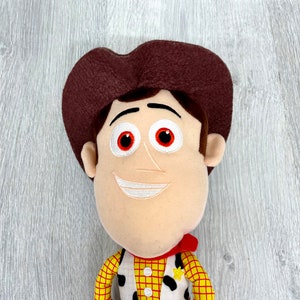 May include: A plush toy of Woody from the Toy Story franchise. He is wearing his signature cowboy hat, vest, and red bandana. He has a friendly smile and red eyes.