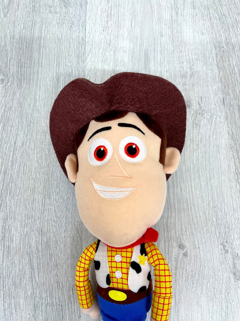 May include: A plush toy of Woody from the Toy Story franchise. He is wearing his signature cowboy hat, vest, and red bandana. He has a friendly smile and red eyes.