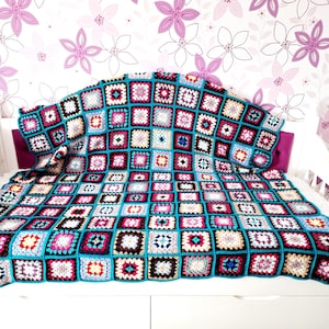 May include: A vibrant, hand-crocheted blanket with a square design. The blanket has a turquoise border and squares in shades of pink, white, black, and brown. The blanket is draped over a white cot.