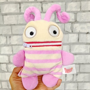May include: A plush toy in the shape of a cartoon alien. The alien has a cream-colored face with large black eyes, pink antennae, and a pink and white striped body. A red zipper runs across the middle. The tag reads "Sorgen Fresser".