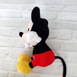 May include: A plush Mickey Mouse toy sitting on a white brick background. The toy is black with red shorts and yellow shoes. The toy has a long black tail.