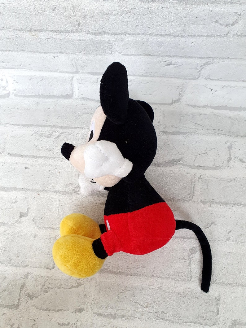 May include: A plush Mickey Mouse toy sitting on a white brick background. The toy is black with red shorts and yellow shoes. The toy has a long black tail.
