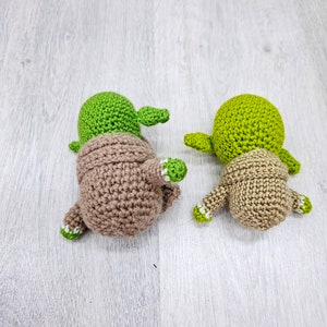 May include: Two crocheted Yoda figures, one green and one brown, with white accents. The figures are facing away from the camera.