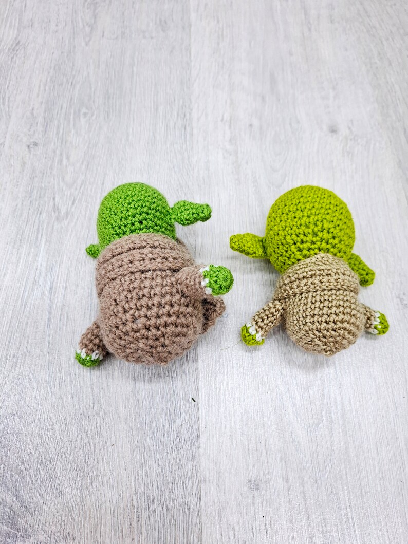 May include: Two crocheted Yoda figures, one green and one brown, with white accents. The figures are facing away from the camera.