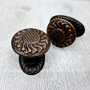 Puede incluir: Dos pomos redondos para armarios con acabado en cobre oscuro. Cada pomo presenta un patrón de rejilla central rodeado por una serie de crestas curvas y radiantes. La base de cada pomo es también circular con una textura de cobre oscuro.
